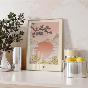 Puede incluir: Una impresi&oacute;n enmarcada de una ilustraci&oacute;n de estilo japon&eacute;s que representa una puesta de sol sobre una masa de agua con una rama de &aacute;rbol con flores en primer plano. La impresi&oacute;n tiene una gama de colores rosa y melocot&oacute;n con flores amarillas en la esquina inferior derecha.