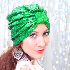 以下が含まれることがあります： 白いトップスと大きな銀のイヤリングをつけた女性が、緑色のスパンコールのターバンをかぶっています。