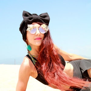 Può includere: Una donna con lunghi capelli rossi che indossa un turbante nero con un fiocco, occhiali da sole a specchio con decorazioni floreali e orecchini turchesi. È sdraiata su una spiaggia sabbiosa.
