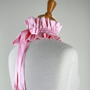 Puede incluir: Un collar de satén rosa con un cuello de volantes y lazos largos. El collar se muestra en un maniquí blanco.