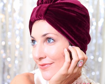 Puede incluir: Una mujer con un turbante de terciopelo burdeos, una blusa blanca con ribete de encaje y varias pulseras de colores. Lleva un anillo con una piedra rosa y tiene una flor rosa prendida en la blusa.