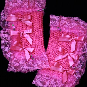 Puede incluir: Un par de calentadores de mu&ntilde;eca de crochet rosa con ribete de encaje y lazos de sat&eacute;n rosa. Los calentadores tienen peque&ntilde;os adornos de flores rosas.