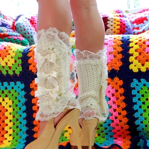 Puede incluir: Un par de polainas blancas de ganchillo con ribete de encaje y lazos, usadas con tacones de plataforma beige. Los tacones est&aacute;n sobre una manta de ganchillo de colores.