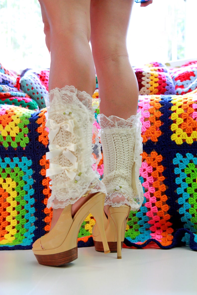 Puede incluir: Un par de polainas blancas de ganchillo con ribete de encaje y lazos, usadas con tacones de plataforma beige. Los tacones est&aacute;n sobre una manta de ganchillo de colores.