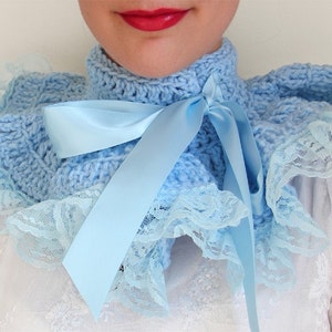 May include: A light blue crocheted collar featuring a high neck design with a matching satin ribbon tied in a bow at the front. The edges of the collar are trimmed with a delicate, ruffled light blue lace fabric that creates a layered effect.