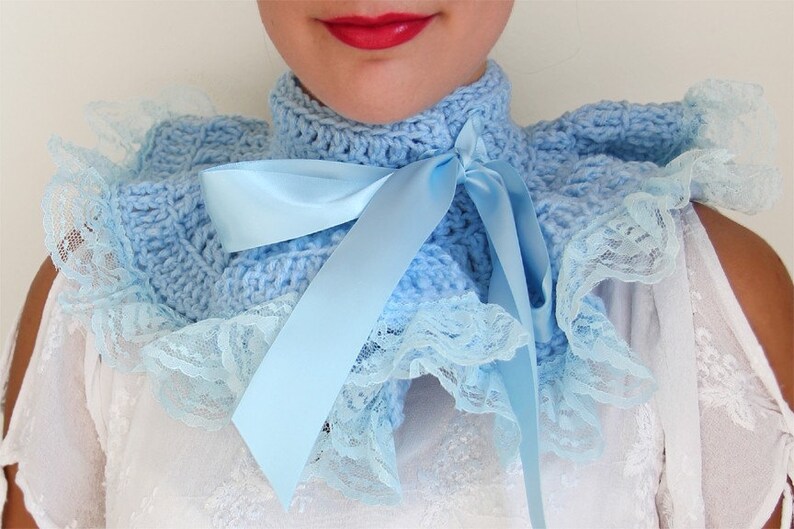 May include: A light blue crocheted collar featuring a high neck design with a matching satin ribbon tied in a bow at the front. The edges of the collar are trimmed with a delicate, ruffled light blue lace fabric that creates a layered effect.