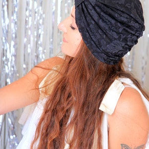 May include: A woman with long, wavy brown hair is wearing a black lace turban. She is wearing a white sleeveless top and has a butterfly tattoo on her upper arm.