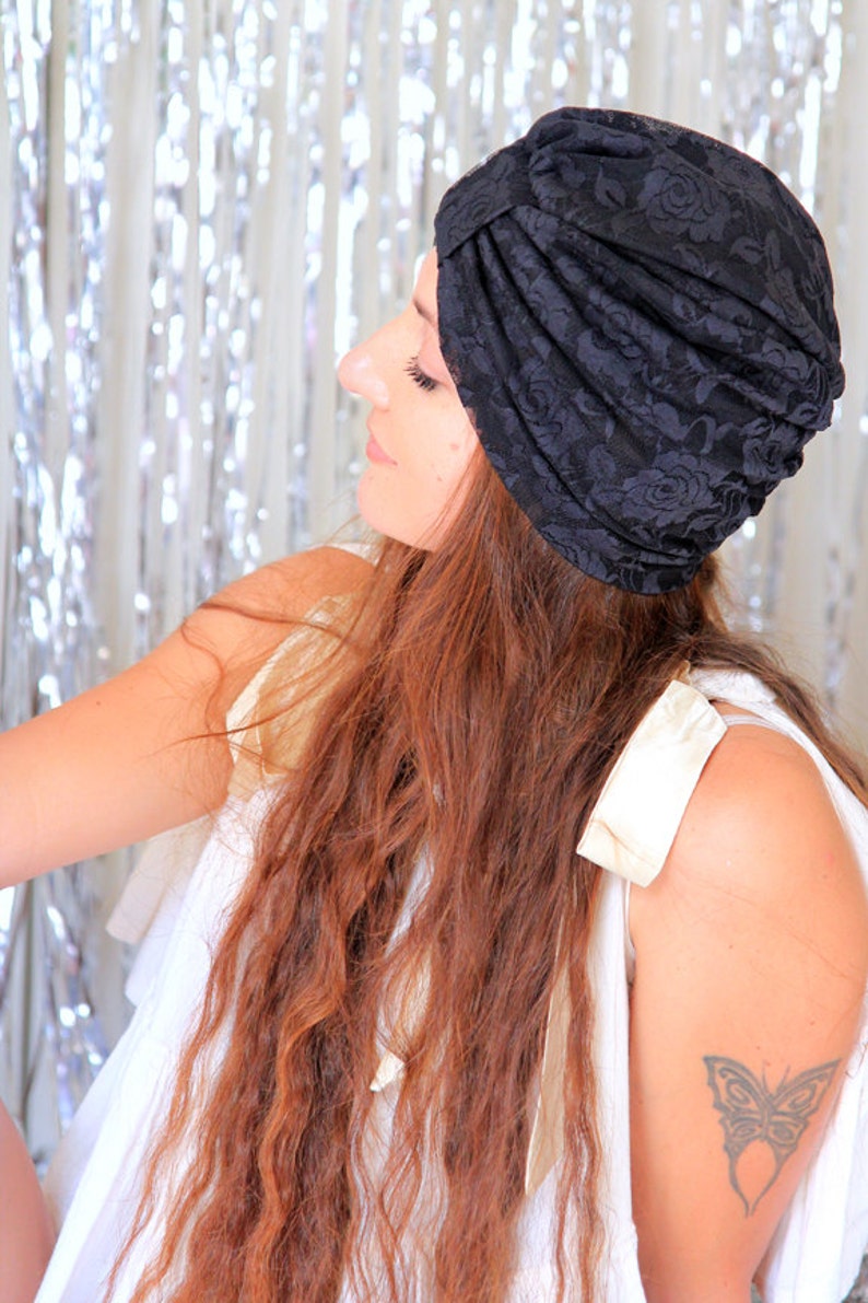 May include: A woman with long, wavy brown hair is wearing a black lace turban. She is wearing a white sleeveless top and has a butterfly tattoo on her upper arm.