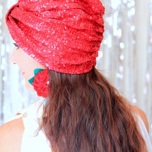 Puede incluir: Un turbante rojo de lentejuelas con un acento de flor roja. El turbante lo lleva una persona con el pelo largo y casta&ntilde;o.