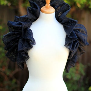 May include: A black lace shoulder wrap with ruffled sleeves is displayed on a white mannequin. The wrap has a high, ruffled collar and is made of a delicate, patterned lace fabric. The background is blurred with green foliage.