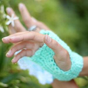 May include: A close-up of a hand wearing a light green crocheted fingerless glove. The hand is reaching out towards a cluster of white flowers.