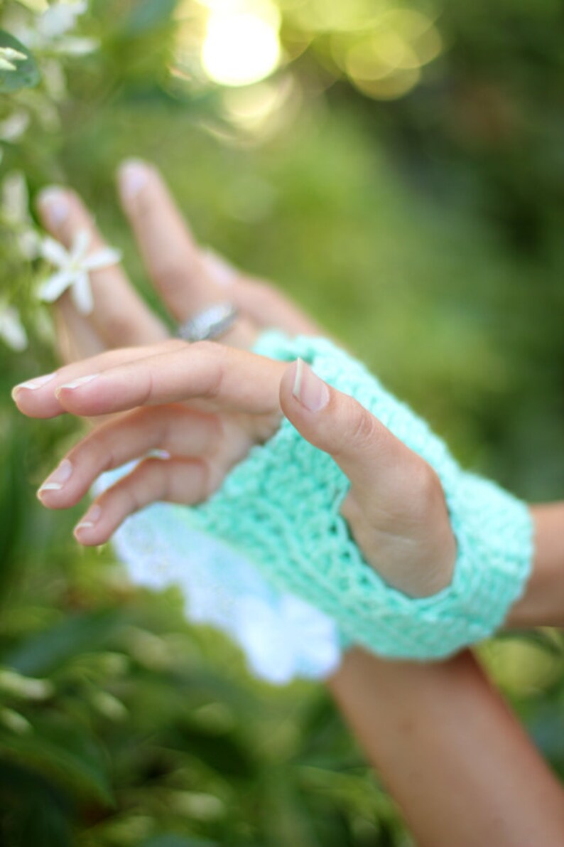 May include: A close-up of a hand wearing a light green crocheted fingerless glove. The hand is reaching out towards a cluster of white flowers.