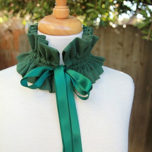 May include: A green fabric choker with a ruffled edge and a matching green satin ribbon tied in a bow. The choker is displayed on a white mannequin.