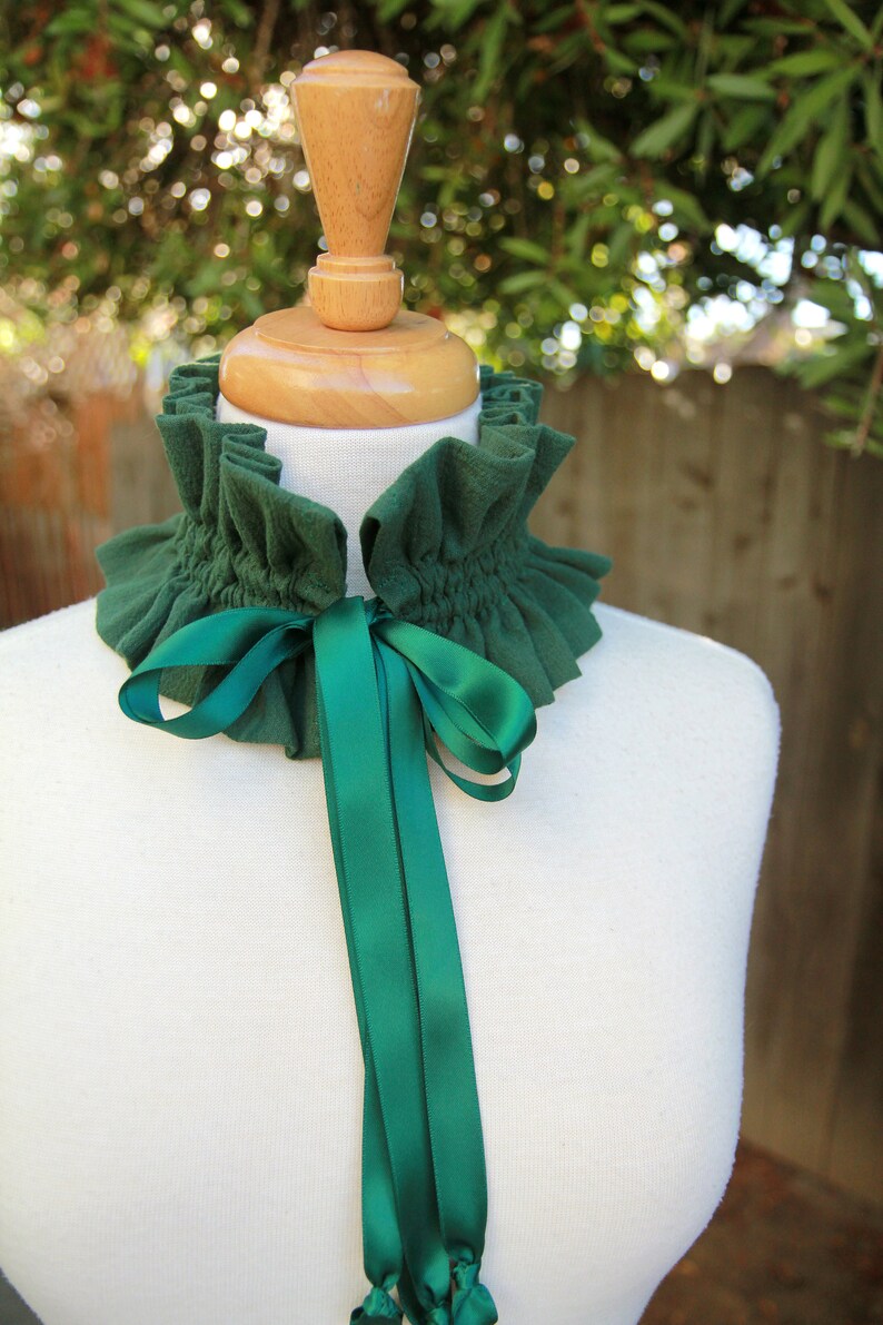 May include: A green fabric choker with a ruffled edge and a matching green satin ribbon tied in a bow. The choker is displayed on a white mannequin.