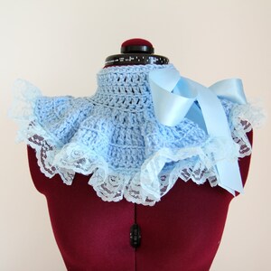 May include: A light blue crocheted collar featuring a high neck design with a ruffled edge. The collar is trimmed with white floral lace and includes a long, smooth light blue satin ribbon tied into a bow on the right side of the garment.