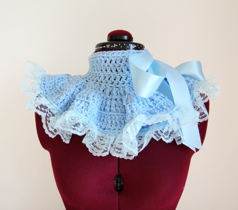 May include: A light blue crocheted collar featuring a high neck design with a ruffled edge. The collar is trimmed with white floral lace and includes a long, smooth light blue satin ribbon tied into a bow on the right side of the garment.