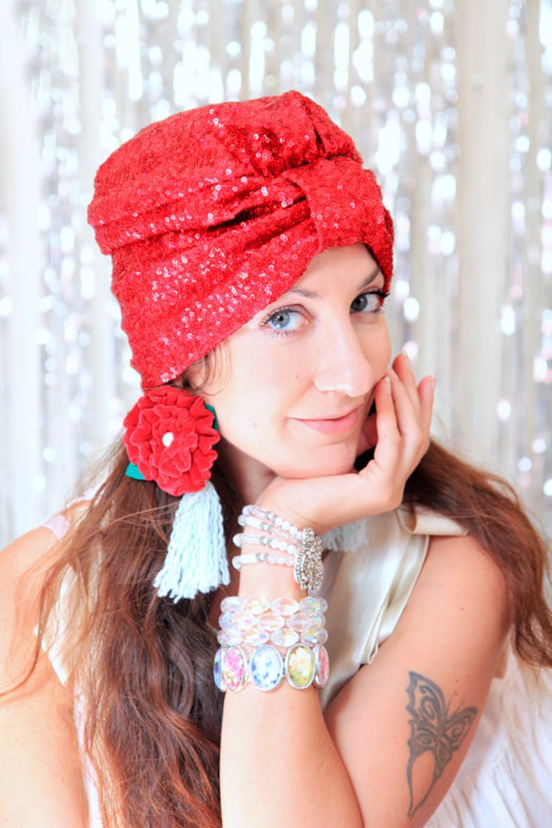 Puede incluir: Una mujer con un turbante rojo de lentejuelas, pendientes de flores rojas y varios brazaletes. Lleva una blusa blanca y tiene un tatuaje de mariposa en el brazo.