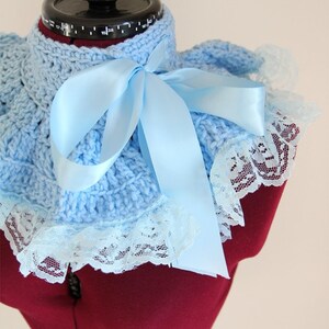 May include: A light blue crocheted collar featuring a textured knit pattern, a wide satin ribbon tied in a bow at the center, and a delicate white lace trim along the outer edge. The collar is displayed on a dark red mannequin torso.