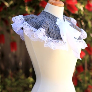 May include: A gray crocheted collar with a white lace trim and a white satin bow. The collar is displayed on a white dress form.