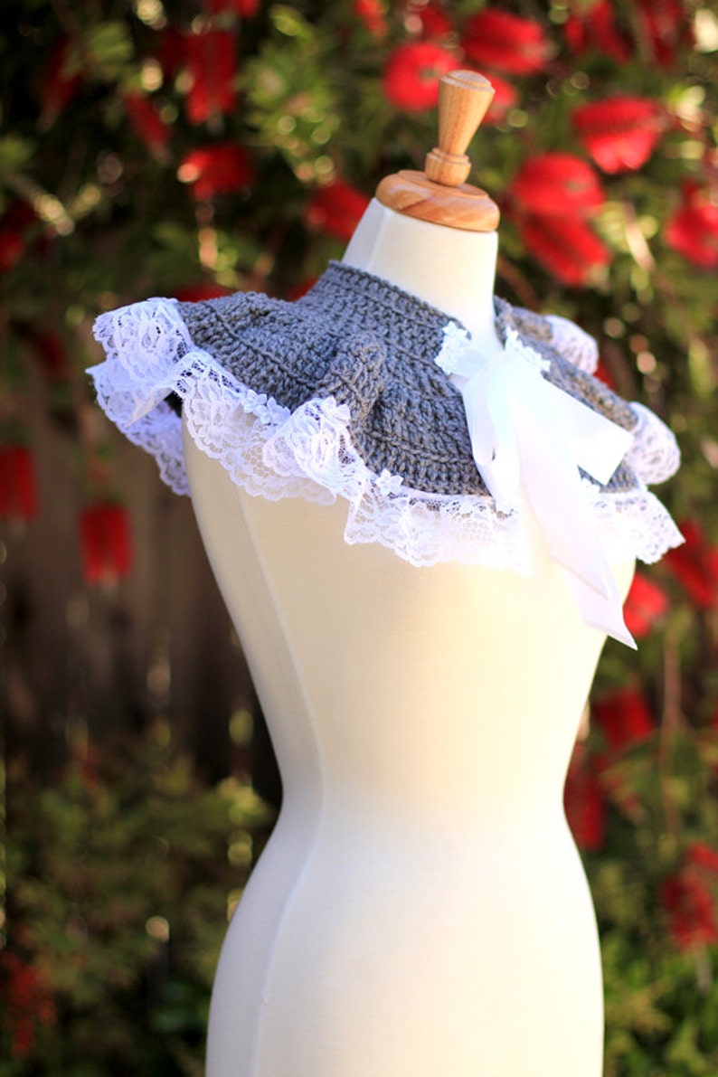 May include: A gray crocheted collar with a white lace trim and a white satin bow. The collar is displayed on a white dress form.