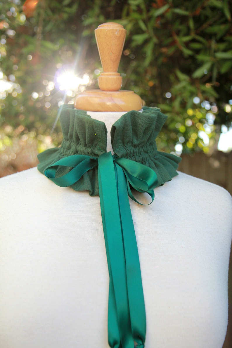 May include: A green ruffled collar with a matching green satin ribbon tied in a bow. The collar is displayed on a white mannequin.