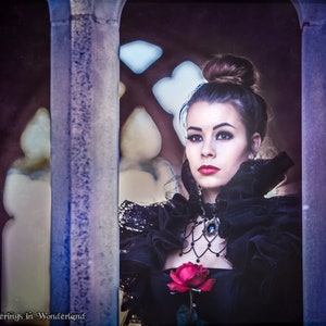 May include: A woman in a black dress with a lace collar and a red rose in her hand looks out of a window. She is wearing a black choker necklace with a jeweled pendant.