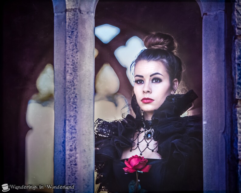 May include: A woman in a black dress with a lace collar and a red rose in her hand looks out of a window. She is wearing a black choker necklace with a jeweled pendant.
