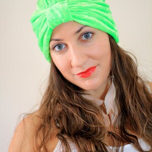 May include: A woman with long brown hair is wearing a bright green turban. She is looking at the camera and has a slight smile on her face. She is wearing a white tank top.