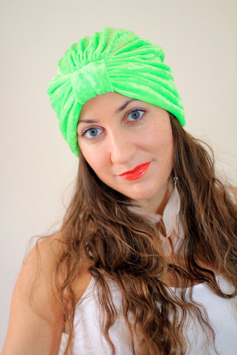 May include: A woman with long brown hair is wearing a bright green turban. She is looking at the camera and has a slight smile on her face. She is wearing a white tank top.