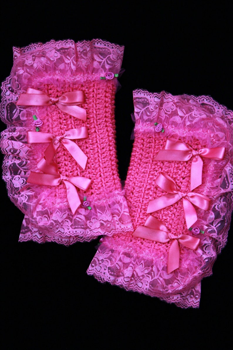 Puede incluir: Un par de calentadores de mu&ntilde;eca de crochet rosa con ribete de encaje y lazos de sat&eacute;n rosa. Los calentadores tienen peque&ntilde;os adornos de flores rosas.