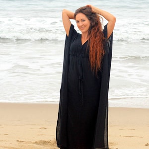 Puede incluir: Una mujer con un vestido largo de caft&aacute;n negro y fluido en una playa de arena. El vestido tiene un escote en V y mangas largas. La mujer est&aacute; de pie con los brazos levantados por encima de la cabeza, y su largo cabello rojo cae por su espalda.