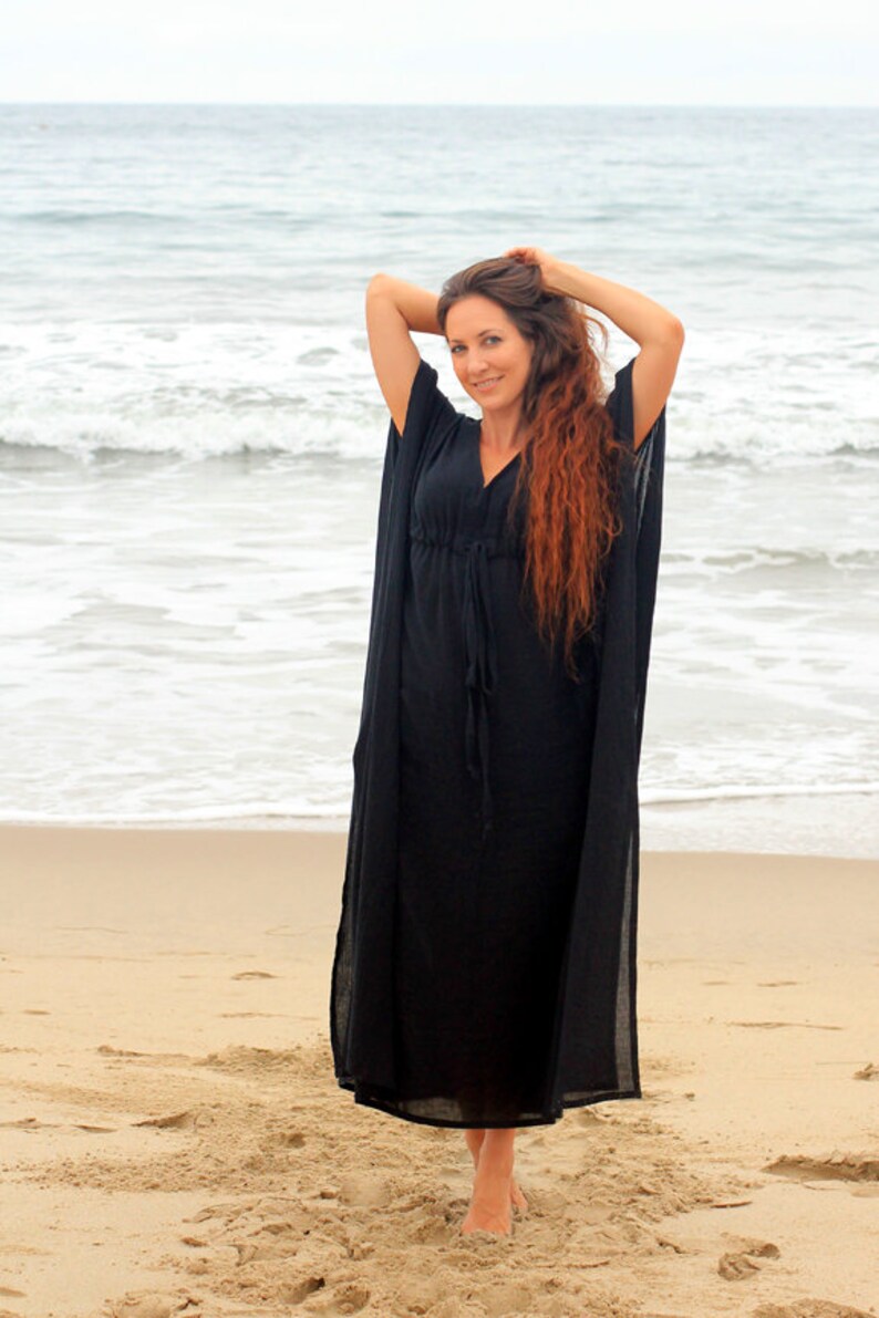 Puede incluir: Una mujer con un vestido largo de caft&aacute;n negro y fluido en una playa de arena. El vestido tiene un escote en V y mangas largas. La mujer est&aacute; de pie con los brazos levantados por encima de la cabeza, y su largo cabello rojo cae por su espalda.
