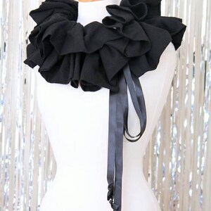 May include: A black ruffled collar with a black satin ribbon tie. The collar is made of fabric and is worn around the neck.