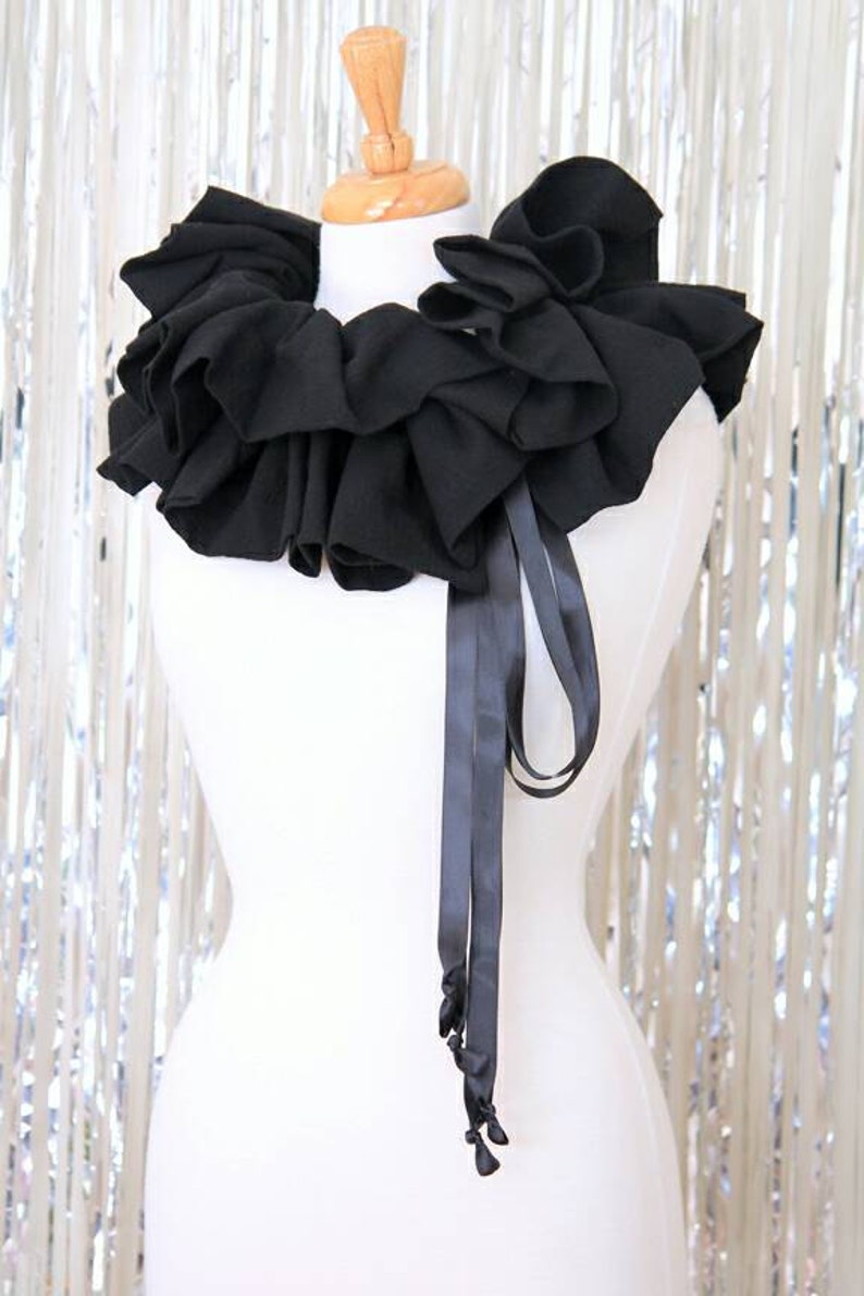 May include: A black ruffled collar with a black satin ribbon tie. The collar is made of fabric and is worn around the neck.