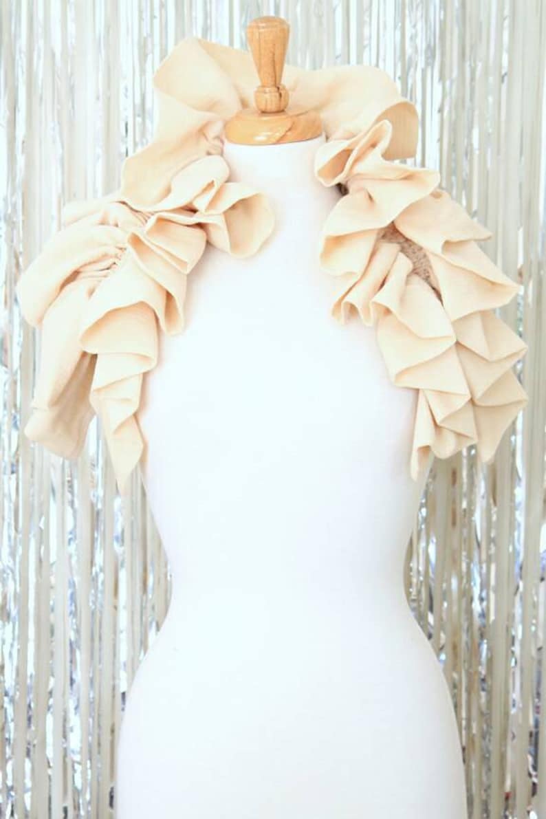 May include: A cream-coloured ruffled fabric collar, worn on a white mannequin. The collar is made of soft, flowing fabric and has a delicate, feminine look.