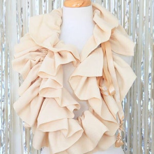 May include: A cream-coloured ruffled scarf with a satin ribbon tie. The scarf is displayed on a white mannequin.