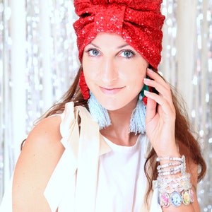 Puede incluir: Una mujer con un turbante rojo de lentejuelas, un top blanco y pendientes de borla azul y blanco. Lleva varias pulseras en la mu&ntilde;eca.