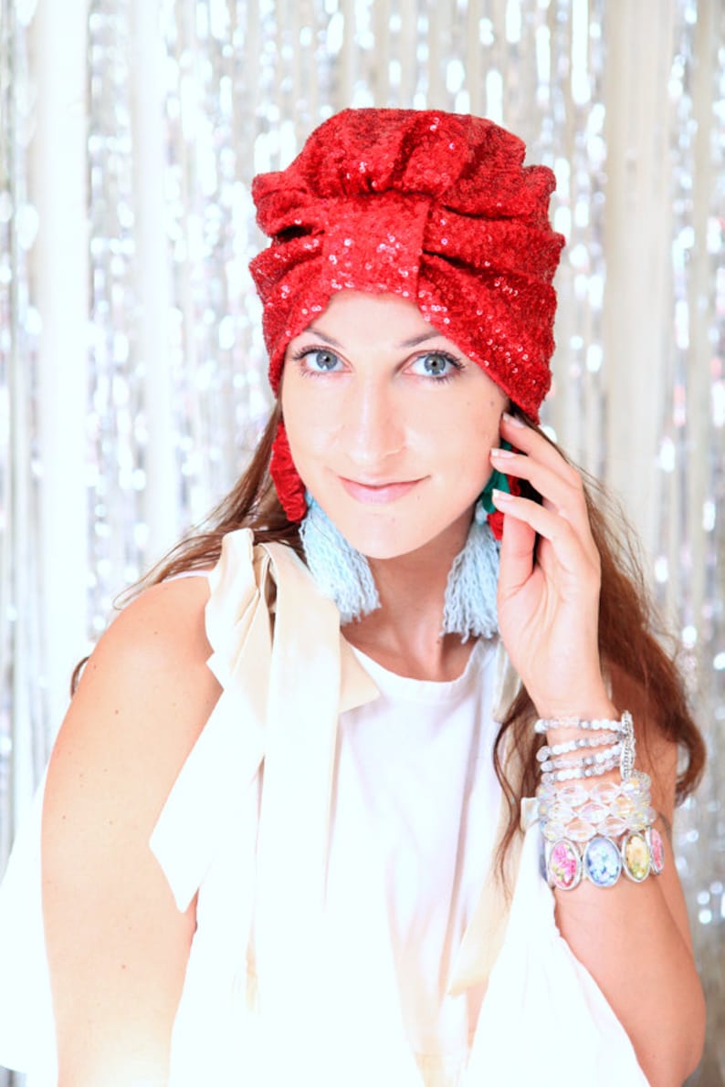 Puede incluir: Una mujer con un turbante rojo de lentejuelas, un top blanco y pendientes de borla azul y blanco. Lleva varias pulseras en la mu&ntilde;eca.