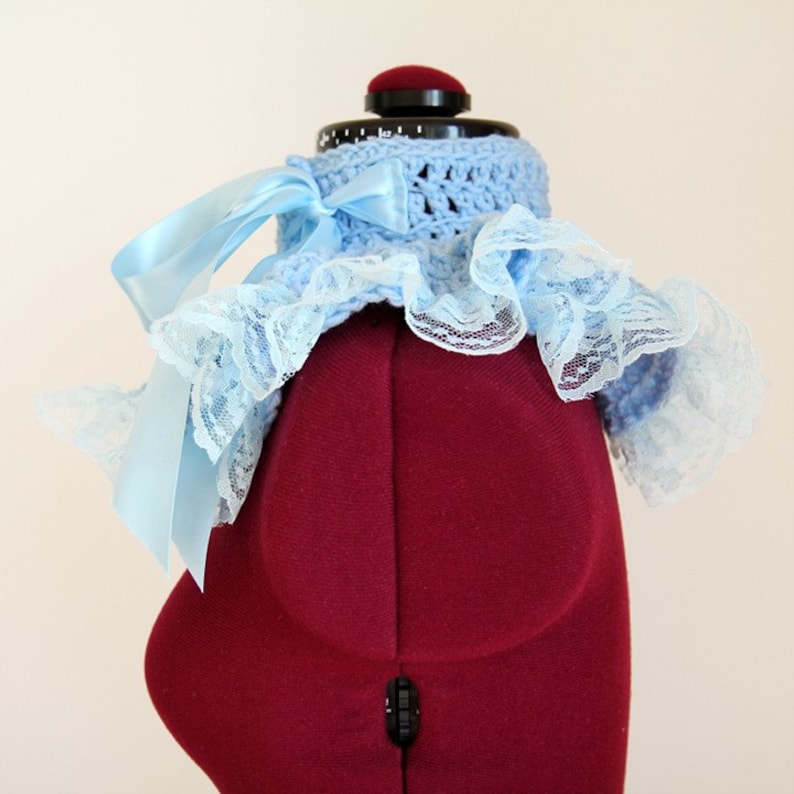 May include: A light blue crocheted collar featuring a wide, ruffled lace trim along the edges and a smooth satin ribbon tied into a bow on the left side. The accessory is displayed on a dark red mannequin torso, highlighting the texture.