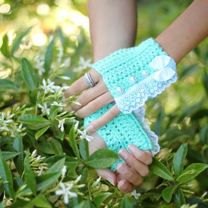 May include: A pair of mint green crocheted fingerless gloves with white lace trim and a white satin bow. The gloves are worn by a person with a ring on their finger.