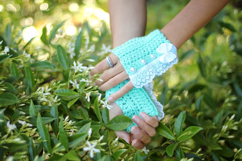 May include: A pair of mint green crocheted fingerless gloves with white lace trim and a white satin bow. The gloves are worn by a person with a ring on their finger.
