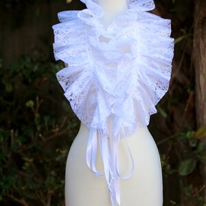 May include: A white lace collar with ruffled edges and satin ribbon ties. The collar is displayed on a white mannequin.