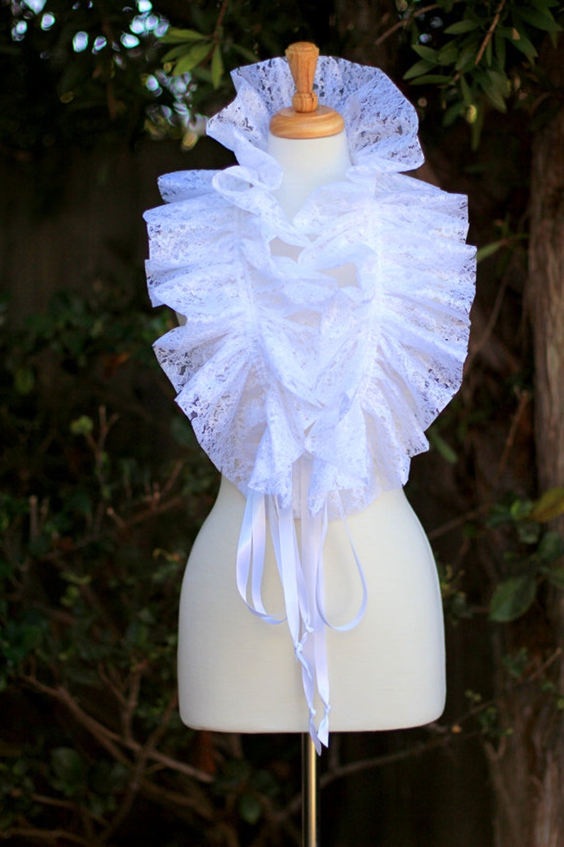 May include: A white lace collar with ruffled edges and satin ribbon ties. The collar is displayed on a white mannequin.