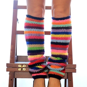 Op de afbeelding: Een paar kleurrijke gestreepte beenwarmers gemaakt van geh&auml;keld garen. De beenwarmers worden gedragen door een persoon die op een houten kruk staat. De beenwarmers zijn een mix van felle kleuren, waaronder rood, oranje, geel, groen, blauw, paars en roze.