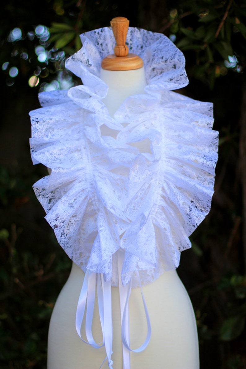 May include: White lace collar with a ruffled design and white satin ribbon ties. The collar is displayed on a white mannequin.