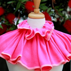 May include: A bright pink ruffled collar with a white trim. The collar is displayed on a white mannequin.