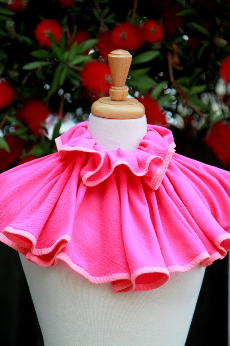 May include: A bright pink ruffled collar with a white trim. The collar is displayed on a white mannequin.
