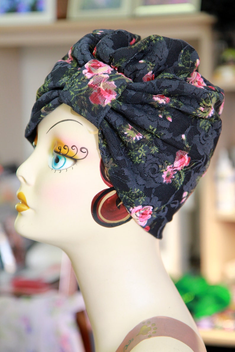 Puede incluir: Un turbante de encaje negro con estampado floral rosa. El turbante se lleva en una cabeza de maniqu&iacute; con maquillaje pintado.