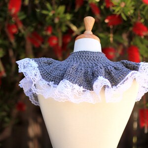 May include: A gray crocheted collar with a white lace trim. The collar is displayed on a white mannequin against a blurred background of red flowers.