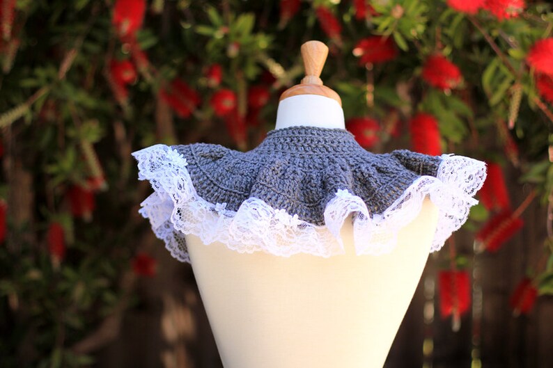May include: A gray crocheted collar with a white lace trim. The collar is displayed on a white mannequin against a blurred background of red flowers.
